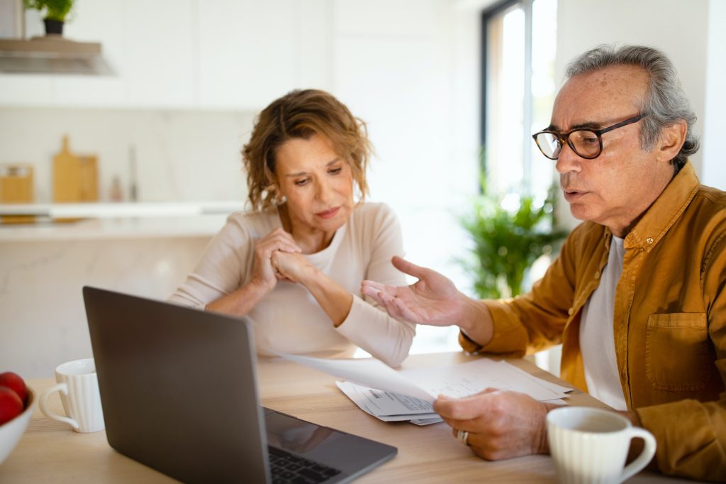 Confused senior husband and wife checking their monthly expenses, reading bills, sitting in front of